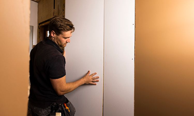 Håndverker i svart T-skjorte setter opp hvit gipsplate i rom under oppussing