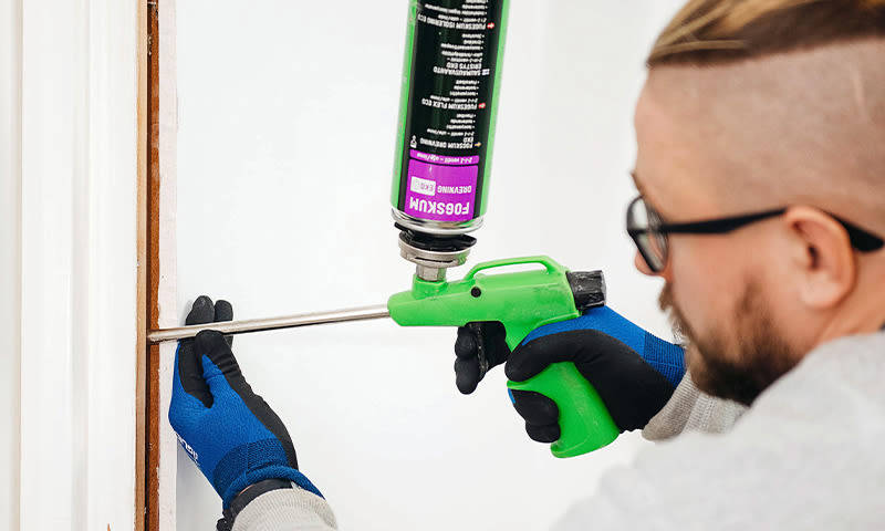 Person med blå hansker bruker grønn skum-pistol for å tette sprekke i hvit dørkarm.
