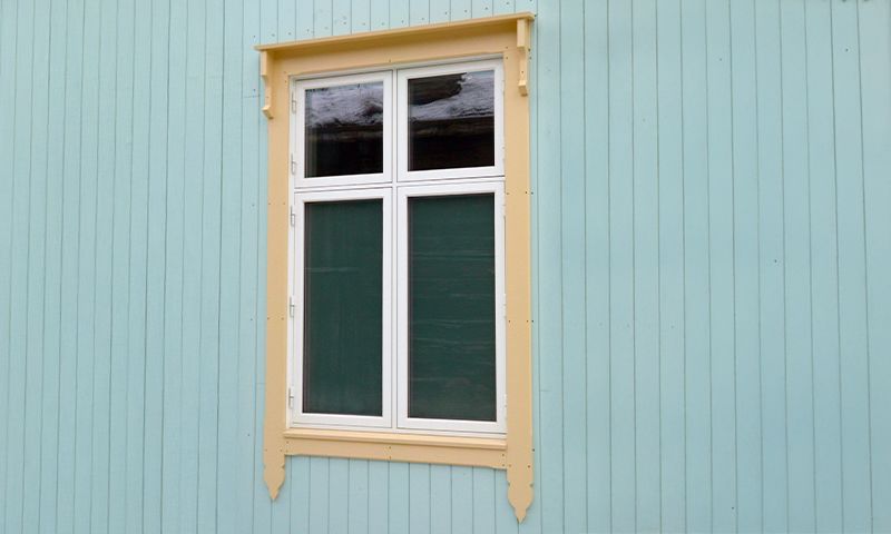 Hvitt vindu med fire ruter, gule lister og grønne lukker på turkis trehusvegg.