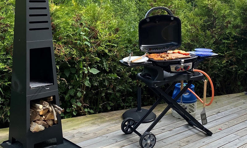 Sort gassgrill på treterrasse med kjøtt og pølser, blå tallerkener, vedstabel og grønn hekk