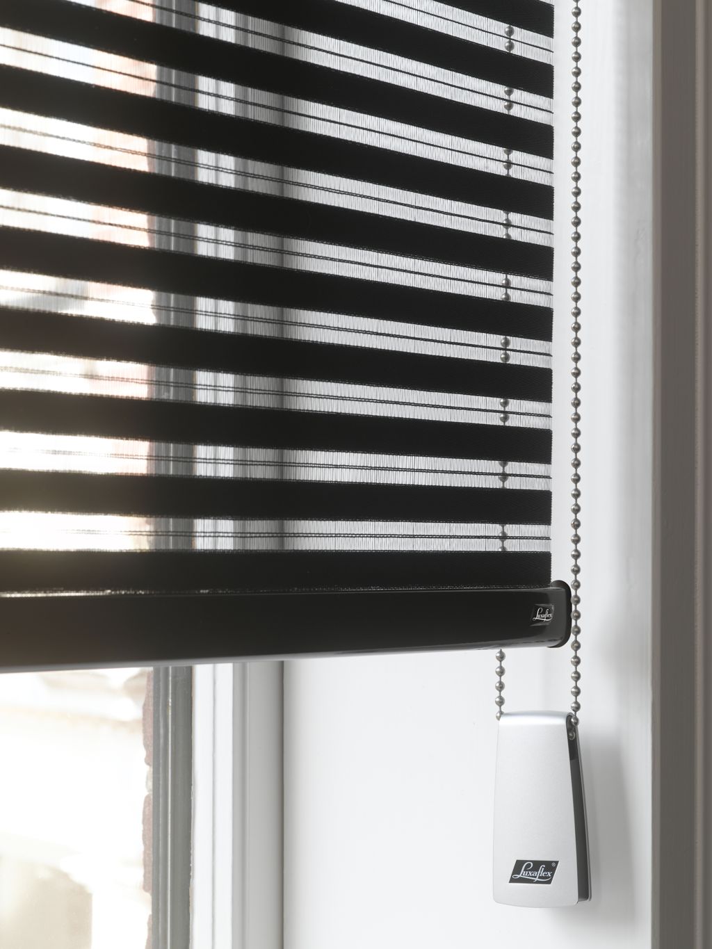 Svart Luxaflex persienne med horisontale lameller og kulekjede foran lyst vindu
