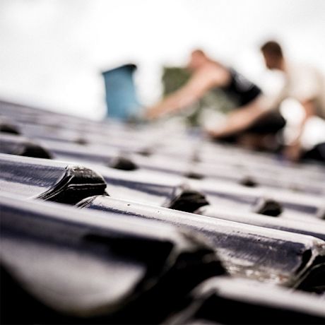 Nærbilde av blanke, mørkeblå takstein mens to håndverkere legger tak i bakgrunnen.