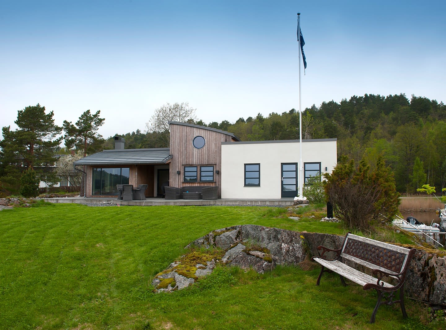 Moderne hvitt og trefasadehus ved vann og skog, grønn plen, flaggstang og benk