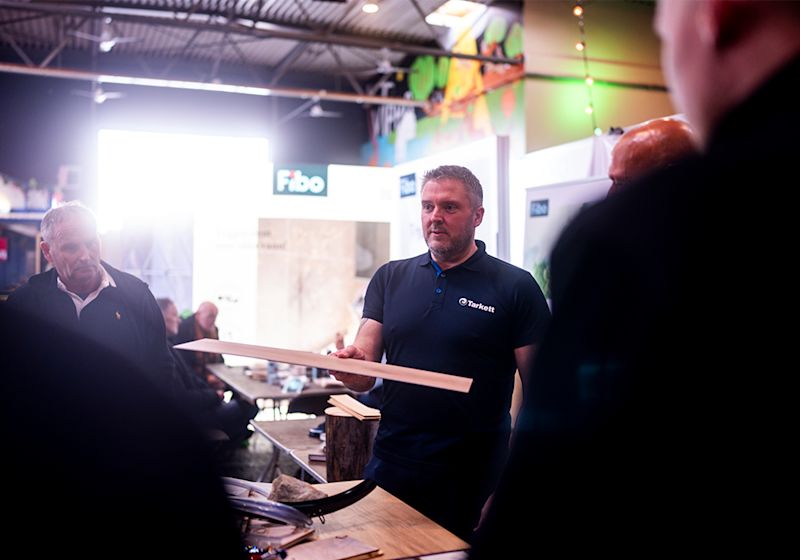 Mann i Tarkett-skjorte viser frem en gulvplanke på en messe med Fibo-stand i bakgrunnen.