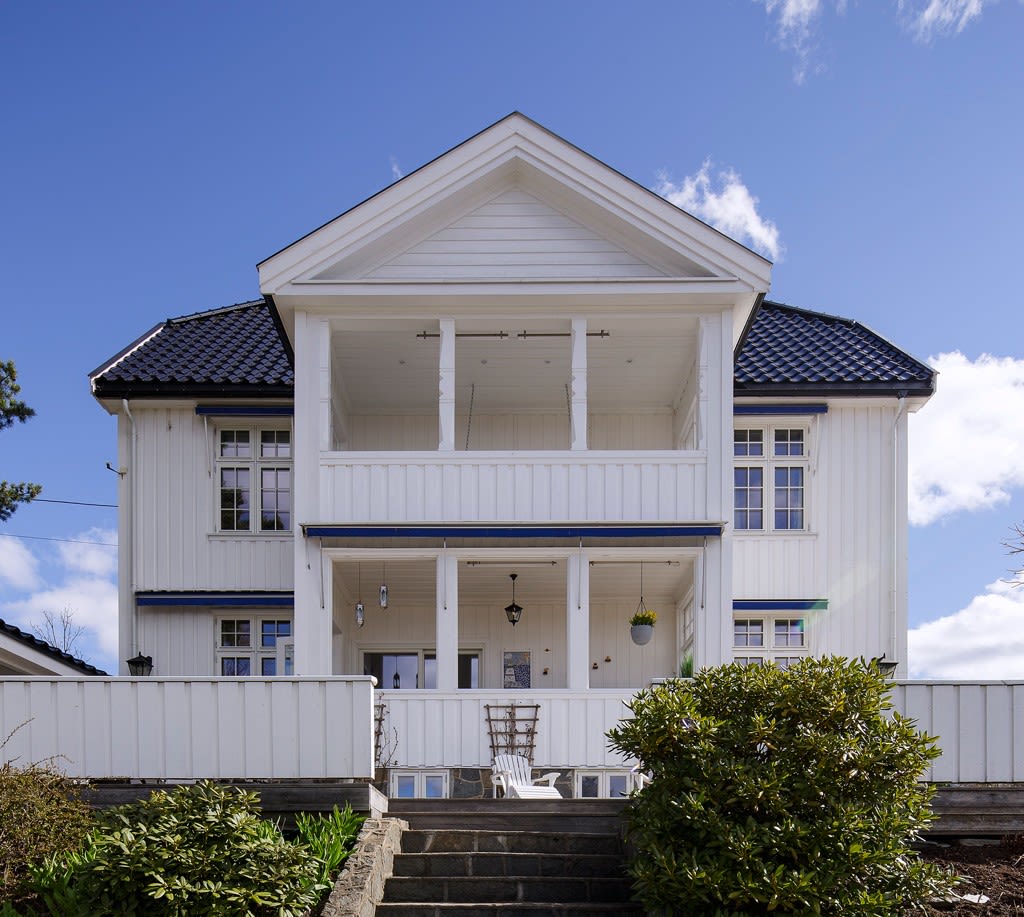 Hvitt toetasjes trehus med blå takstein, veranda og trapp omgitt av grønne busker.