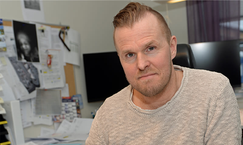 Person i lys beige genser ved overfylt kontorpult med dataskjermer og papirer.