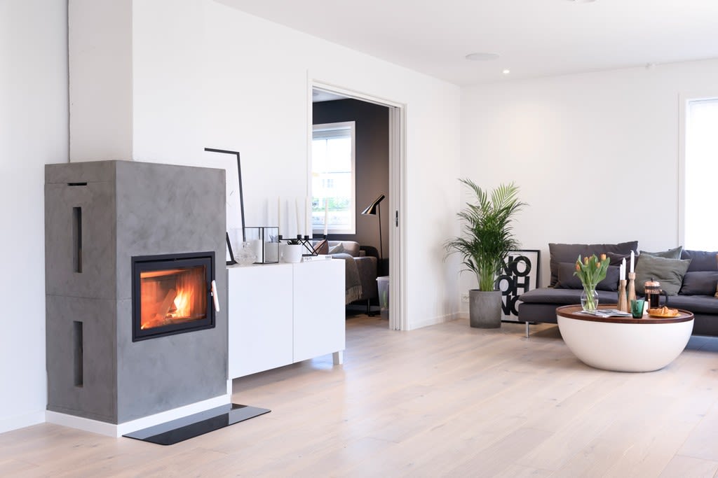 Lyse, minimalistisk stue med grå betongpeis, hvite møbler, grå sofa, grønn plante og rundt bord.