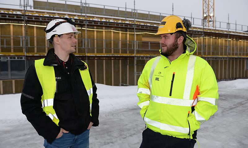 To anleggsarbeidere i gule verneklær og hjelmer ved et snødekt byggeplass med stillas og kran.