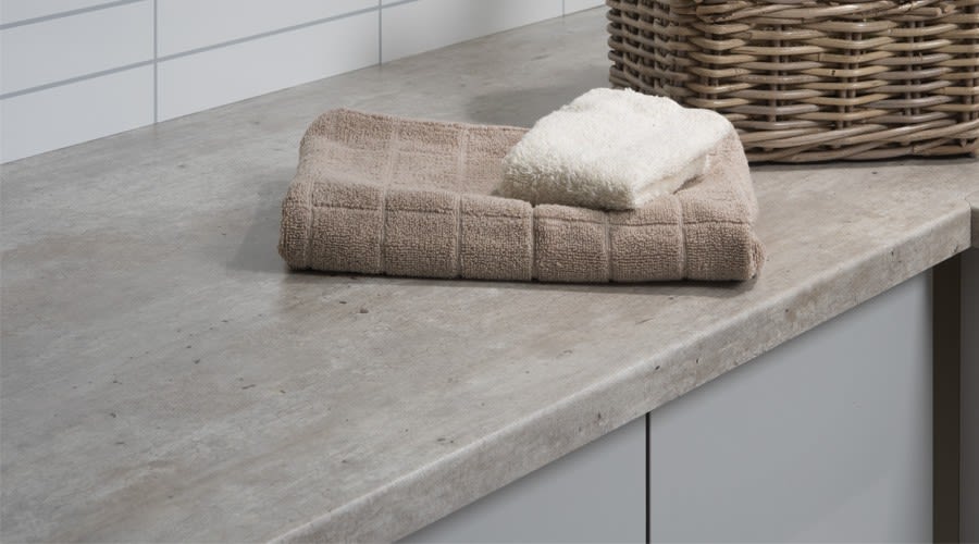 Beige håndkle med hvit klut på grå betongliknende benkeplate ved flettet kurv.