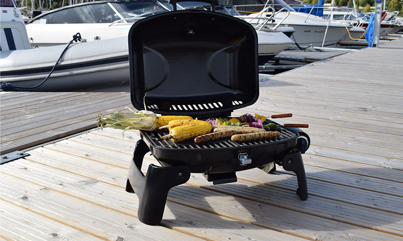 Portabel svart grill med gyllen mais, pølser og grønnsaksspyd på brygge med båter.