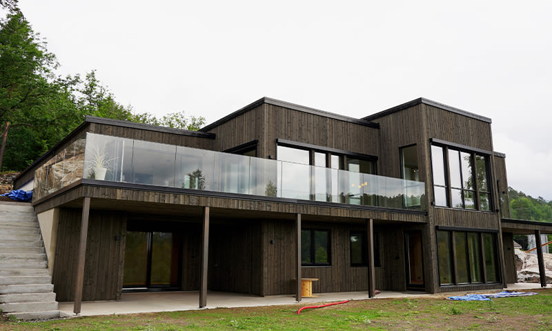 Moderne to-etasjes hus i mørkt treverk, store vinduer og glassrekkverk mot skog.