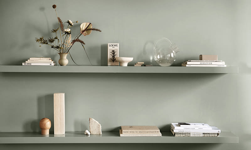 Minimalistiske grønne hyller med lyse bøker, vase med tørkede blomster, bolle og glasskanne.