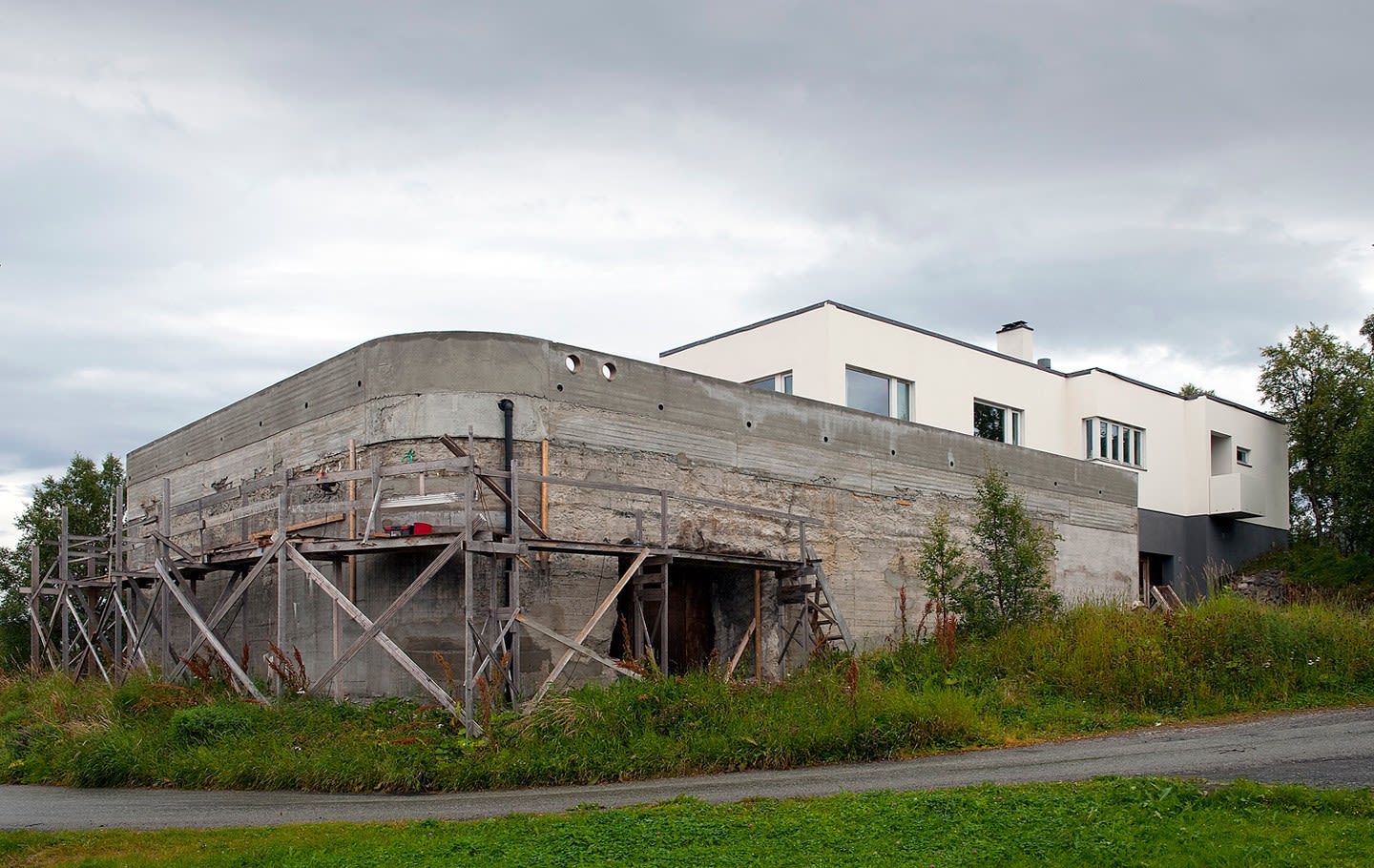 Grå betongbunker under renovering med stillas, moderne hvitt hus bak, gress og skyet himmel.