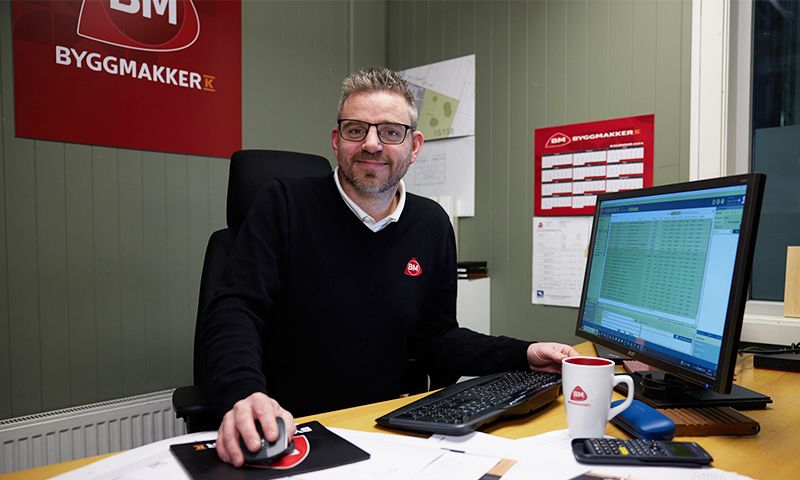 Kontorarbeider i mørk genser ved skrivebord med PC, kaffekopp og rød Byggmakker-logo på grønn vegg.