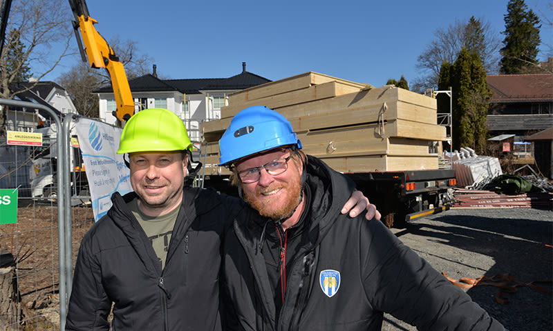 To byggearbeidere med gul og blå hjelm foran lastebil med trepanel på solrik byggeplass.