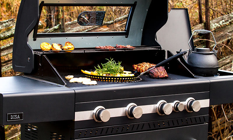 Sort gassgrill i hage med biff, grønnsaker i stekepanne, grillede boller og svart vannkjele.