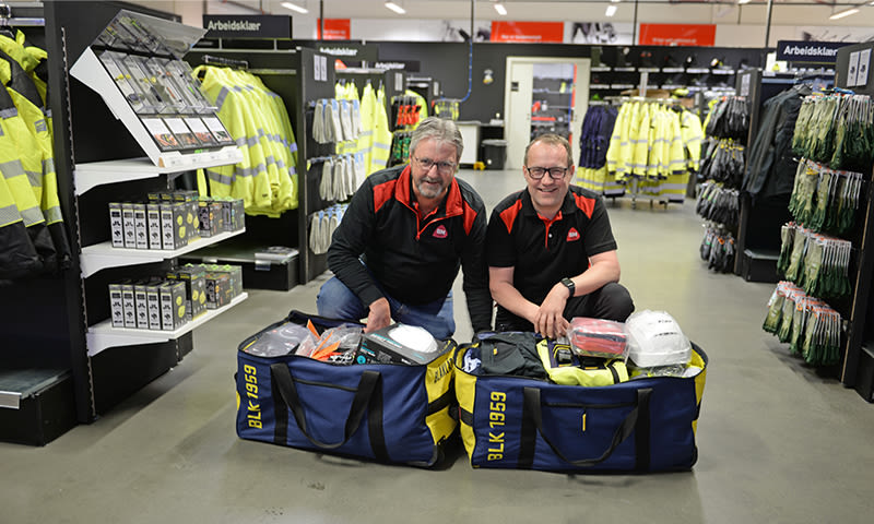 To personer i rødt og svart kneler ved to blågule bagger med verneutstyr i en lys butikk.