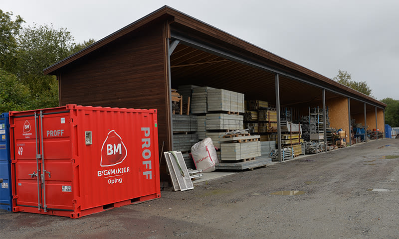 Rød BM Proff-konteiner ved brunt, åpent bygglager med stablede materialer under grå himmel