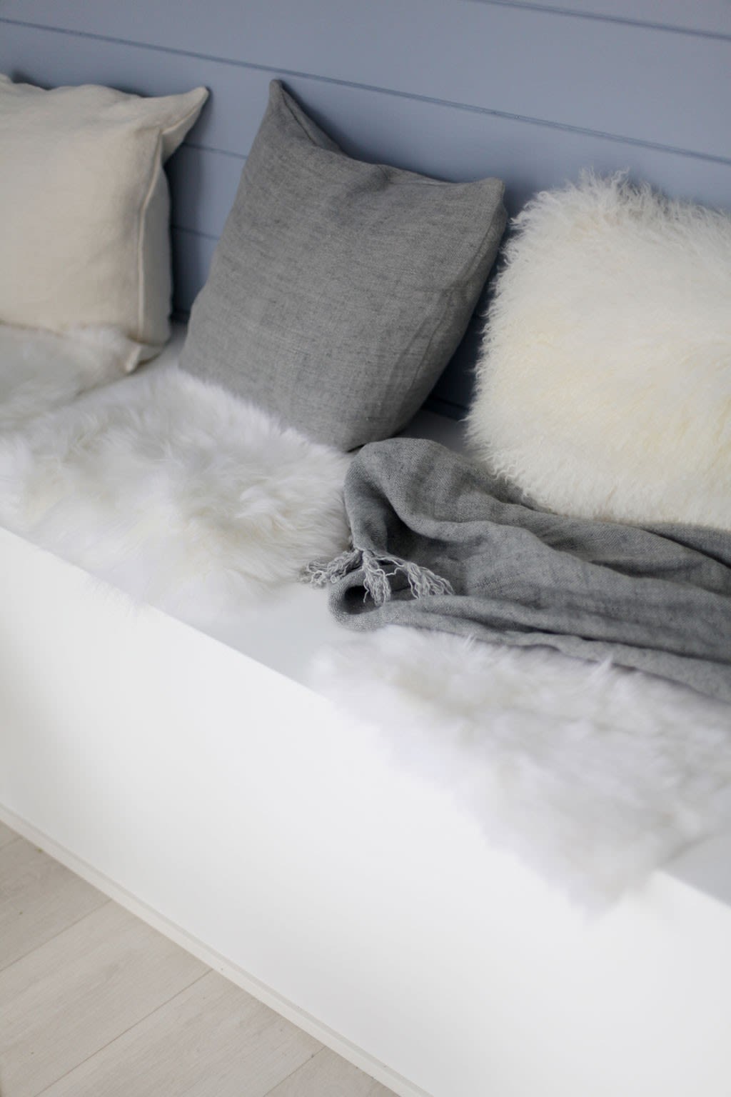 Benken med hvite pelsmatter, grått pledd og puter i krem, grått og hvitt mot lyseblå vegg