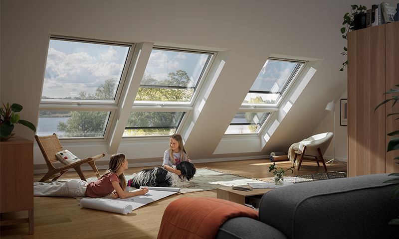 Lyst loftrom med skråvinduer, tregulv, barn som tegner ved hund, moderne møbler og sjøutsikt.