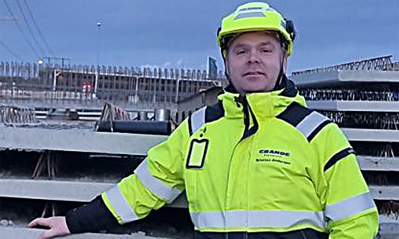 Arbeider i neongul refleksjakke og hjelm foran stabler med armeringsjern på byggeplass.