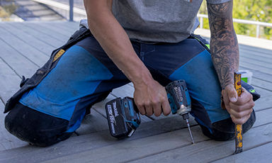 Nærbilde av arbeidshender i blå bukser som borer hull i grå terrasse med batteridrevet drill