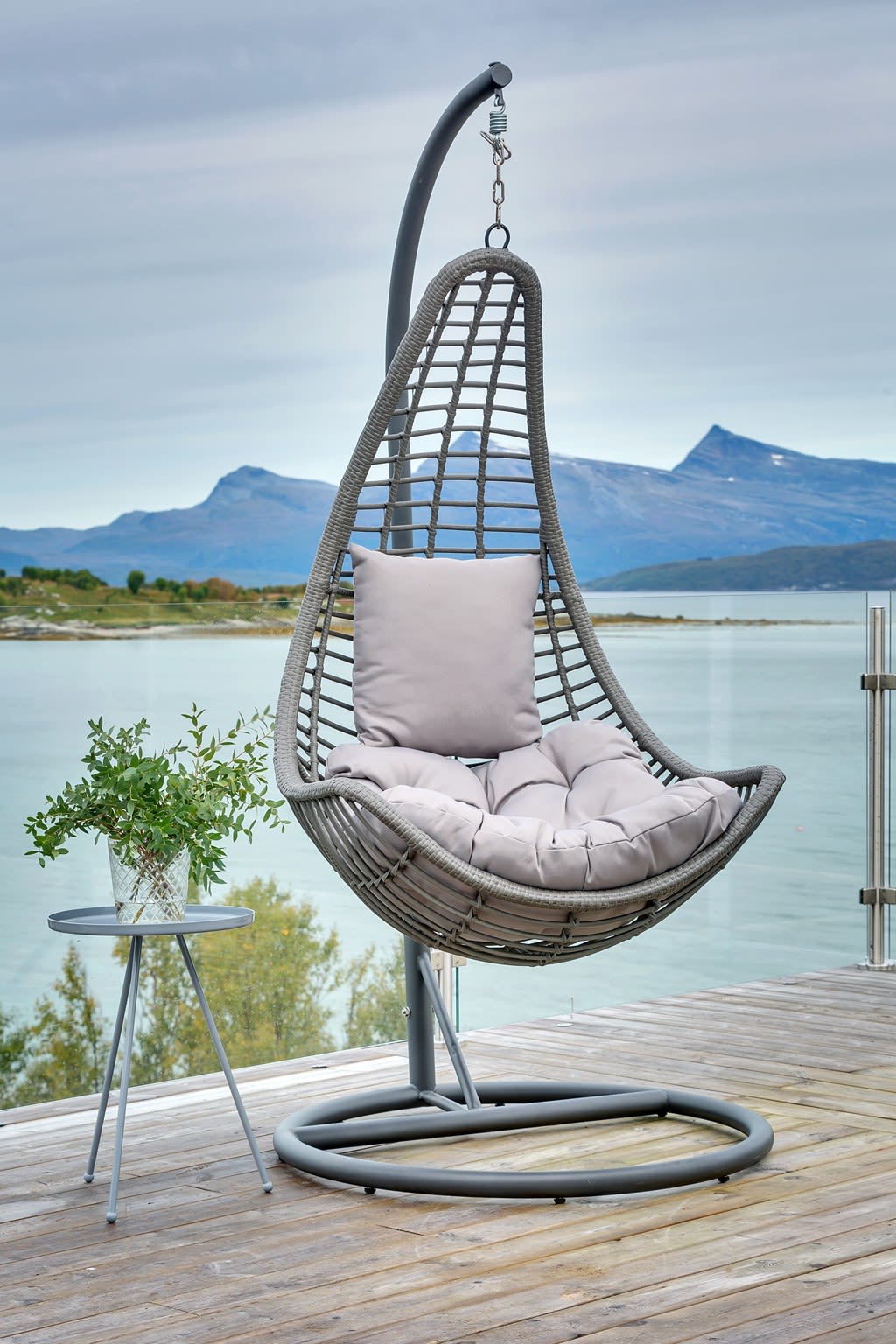 Grå hengestol med lyse puter på terrasse, sidebord med grønn plante og fjordutsikt.