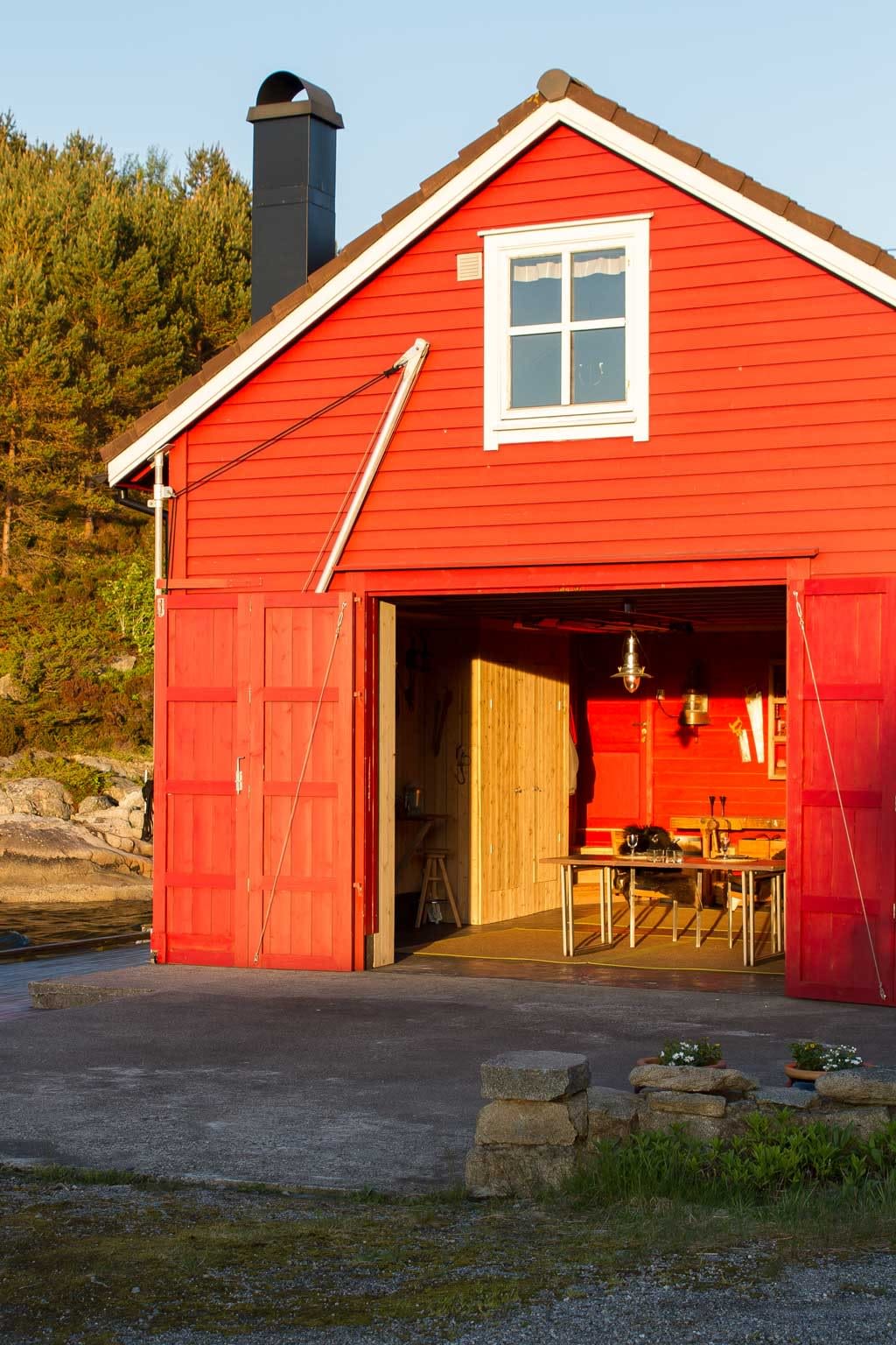 Rødt naust med åpne dobbeltdører ved vann, interiør med bord og lanterne i solnedgang.