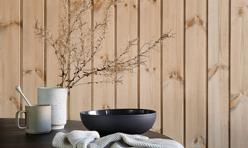 Bord med lys keramikkmugge og -kanne med tørkede strå, svart bolle, strikket klut foran trepanel.