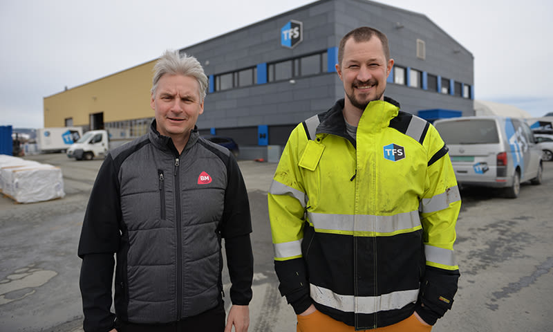 To håndverkere i mørk vest og gul refleksjakke foran grå-gult industribygg med TFS-logo