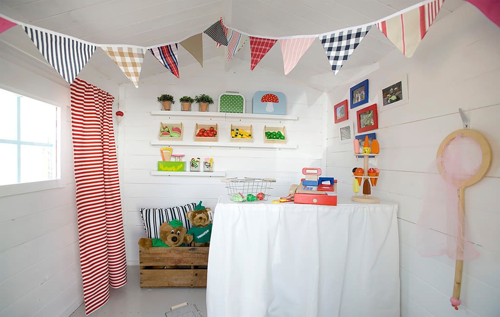 Interiør av hvitt lekeshus med fargerike vimpler, lekemat, kassaapparat og bamser.