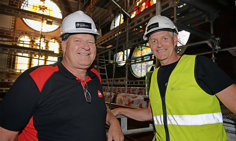 To menn i hvite vernehjelmer ved stillas i kirkebygg med fargerike glassmalerier