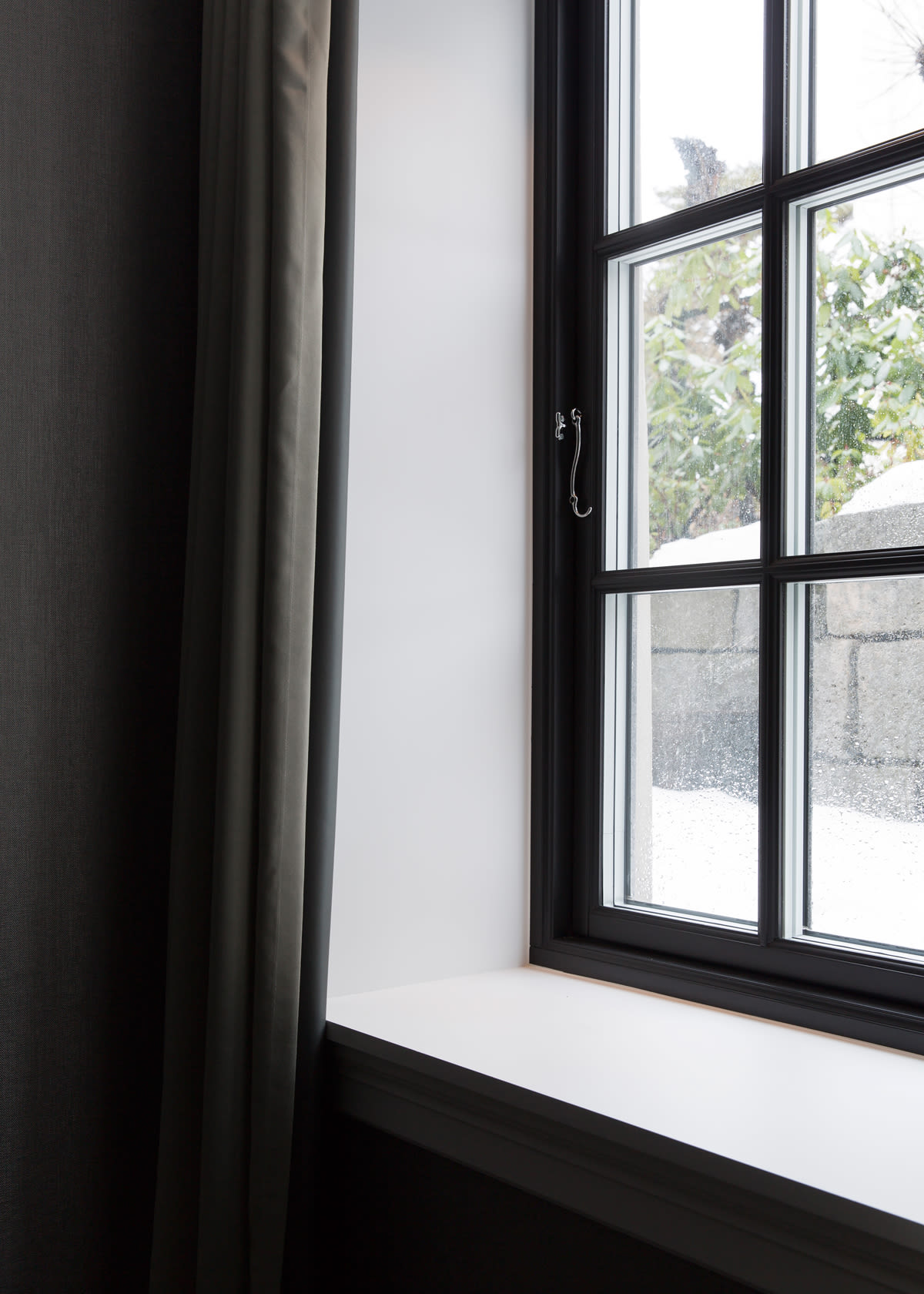 Innvendig vindu med sort ramme, hvit karm og grå gardin mot snødekt steinmur og grønne busker