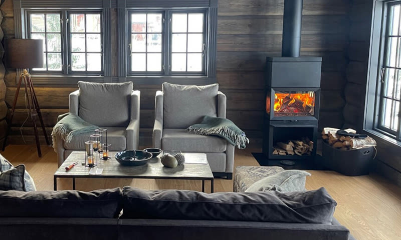 Koselig tømmerstue med to grå lenestoler, dekorativt bord og svart vedovn med ild