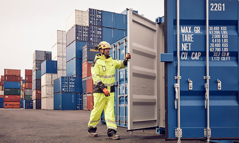 Arbeider i gul vernedrakt og hjelm åpner blå container foran stabler med fargerike containere