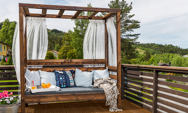 Trepergola med hvite gardiner, blå puter, pledd og brett med juice og appelsiner på grønn terrasse