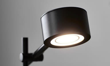 Nærbilde av svart, sylindrisk spotlampe montert på arm med varmt lys mot grå bakgrunn.