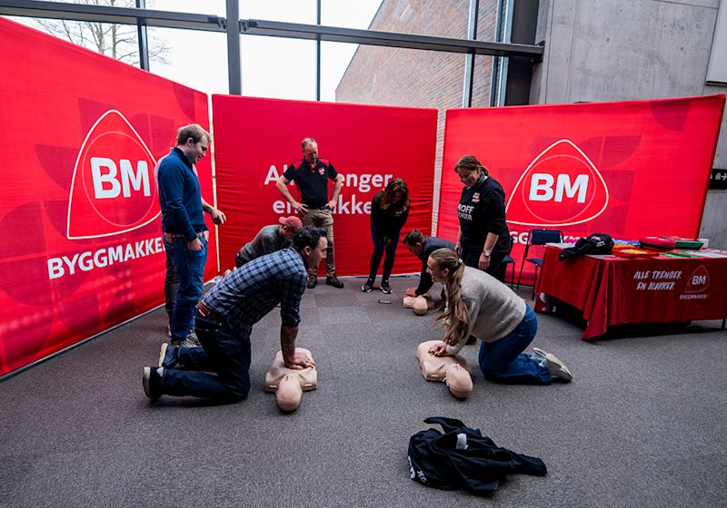 Personer øver hjerte-lunge-redning på dukker foran røde Byggmakker-bannere under et førstehjelpsarrangement.