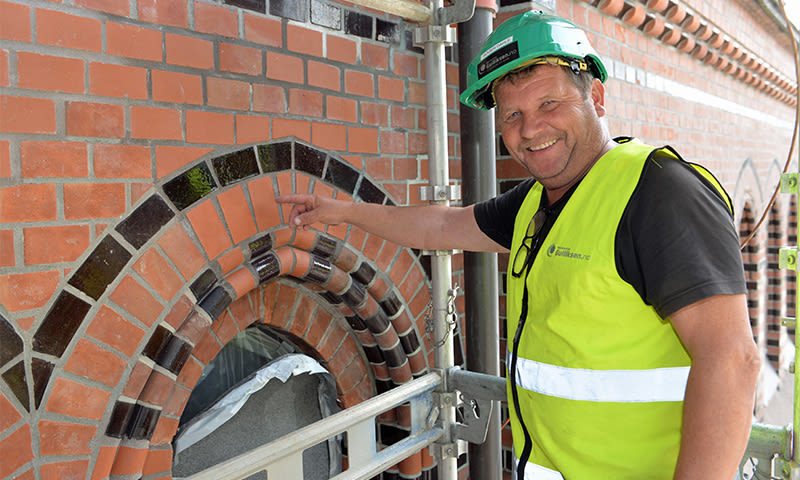 Person i grønn hjelm og gul vest peker på rød mursteinsbue med svarte fliser under stillas