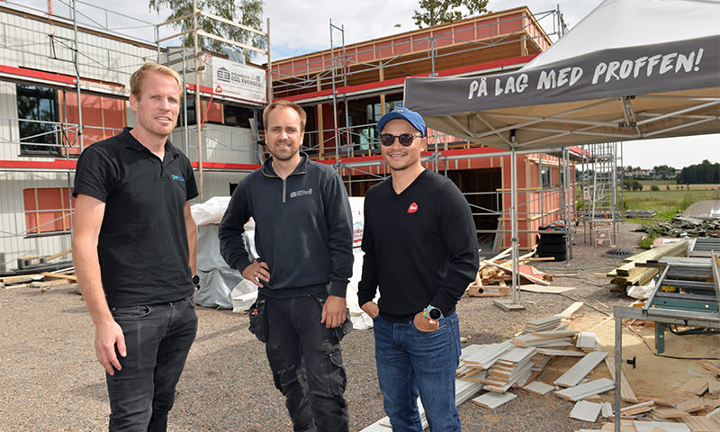 Tre håndverkere i mørke klær foran rød-hvitt bygg under oppføring med stillas og profftelt.