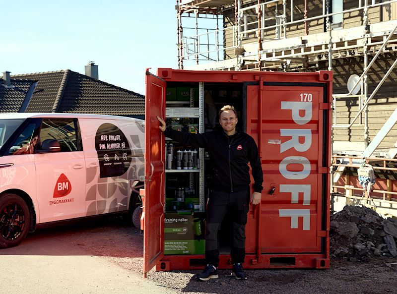 Byggplassen med rødt PROFF-lager, rosa Byggmakker-varebil og montør i svart arbeidstøy.