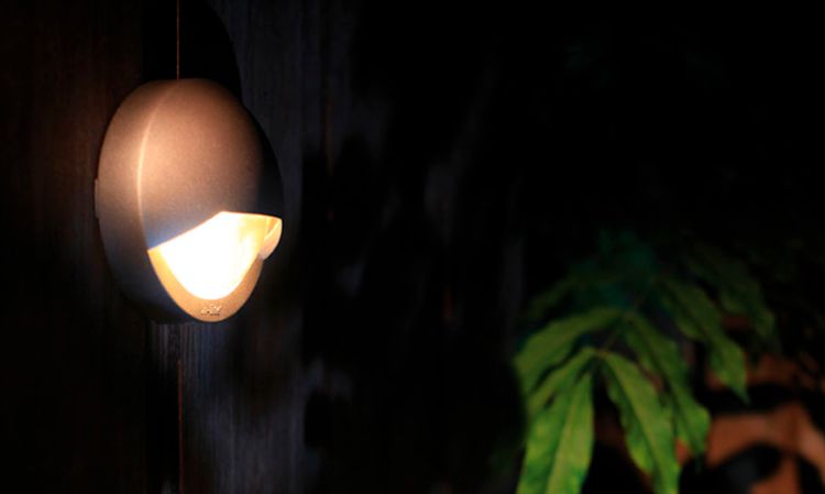 Rund, grå vegglampe med varmt lys retter mot mørkt treverk, grønne blader til høyre.