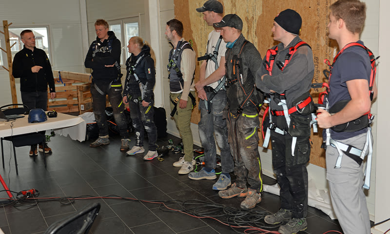 Sju lærlinger i arbeidstøy med svarte fallsikringsseler og røde stropper står inne ved OSB-vegg.