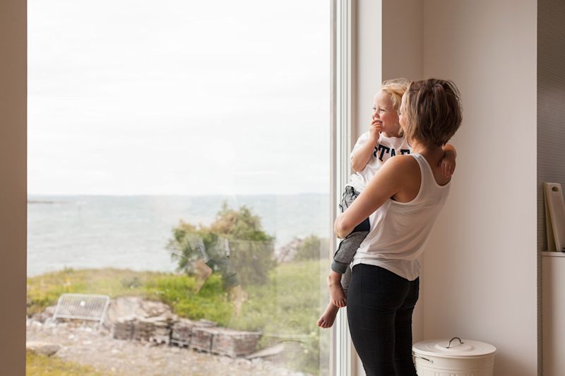 Kvinne i hvit topp holder barn ved stort vindu mot grå sjø og grønne busker.