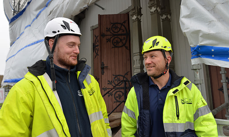 To anleggsarbeidere i gule refleksjakker og hjelmer ved stillas foran dekorativ dør.