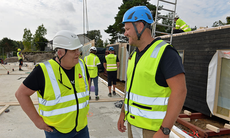 To arbeidere med gule vernevester og hjelmer snakker på byggeplass med stillas og materialer.