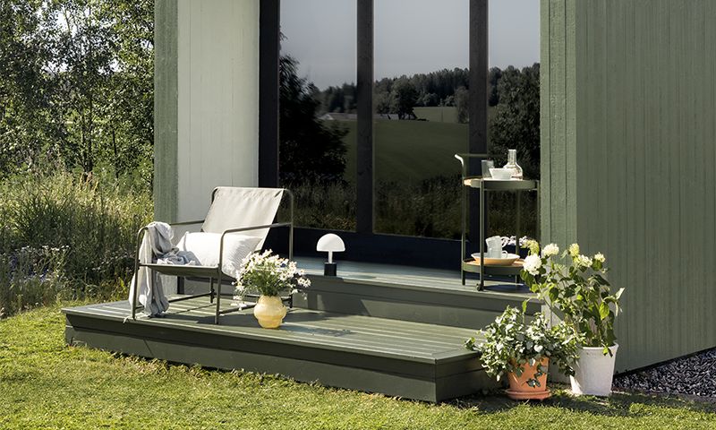 Uteplass ved grønn hytte med lys grå lenestol, gule og hvite blomsterpotter og rullbart bord