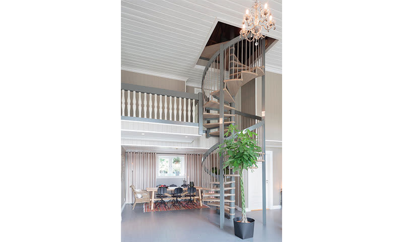 Lyse oppholdsrom med grå spiraltrapp, lysekrone, spisebord med svarte stoler og plante.