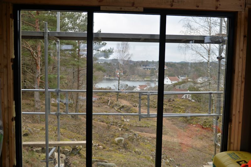 Utsikt gjennom byggeplass-vindu: stillas, furutrær, mosekledt berg, hvite hus ved fjord