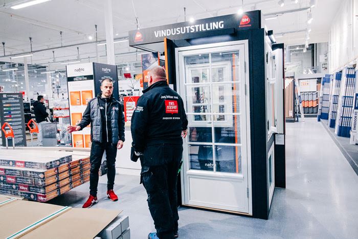 To menn ved sort utstillingsmonter merket «VINDUSUTSTILLING» i lys byggevarebutikk.
